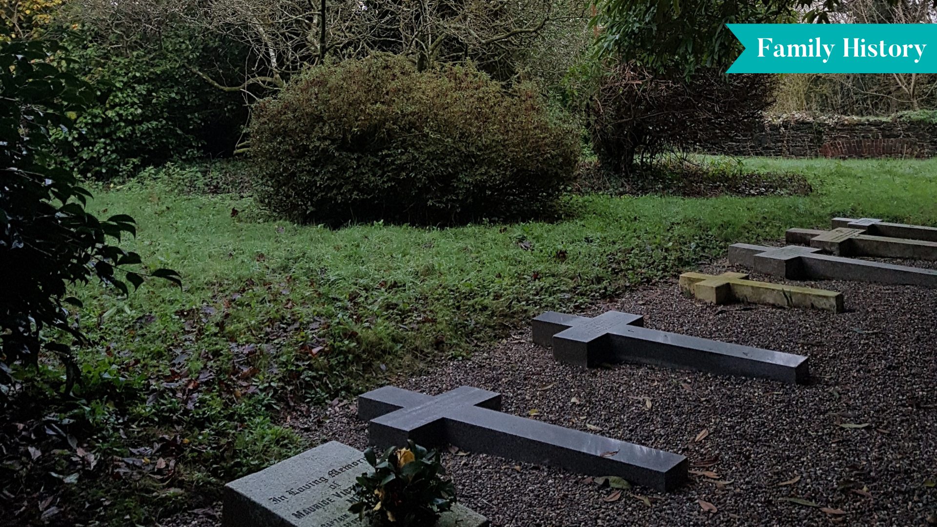 A small family cemetery with six graves arranged in a single neat row along a stone path. Each grave is marked with a simple headstone. The area is surrounded by grass and trees, creating a peaceful and quiet atmosphere under natural daylight.