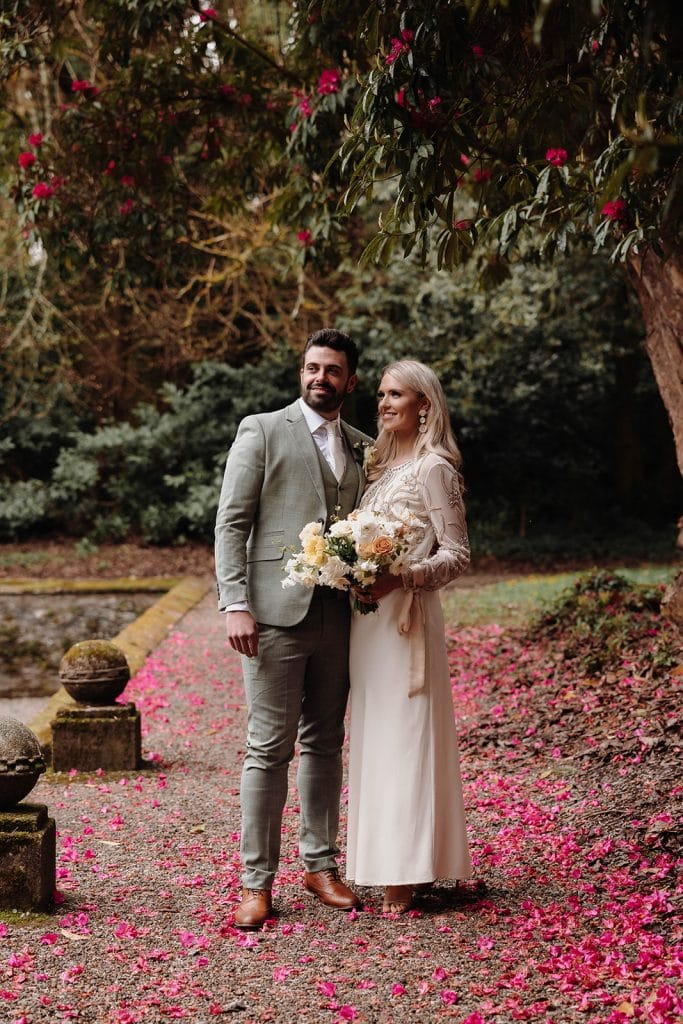 Bride and groom walk pose in picturesque gardens with a carpet of pink petal sunderneaath