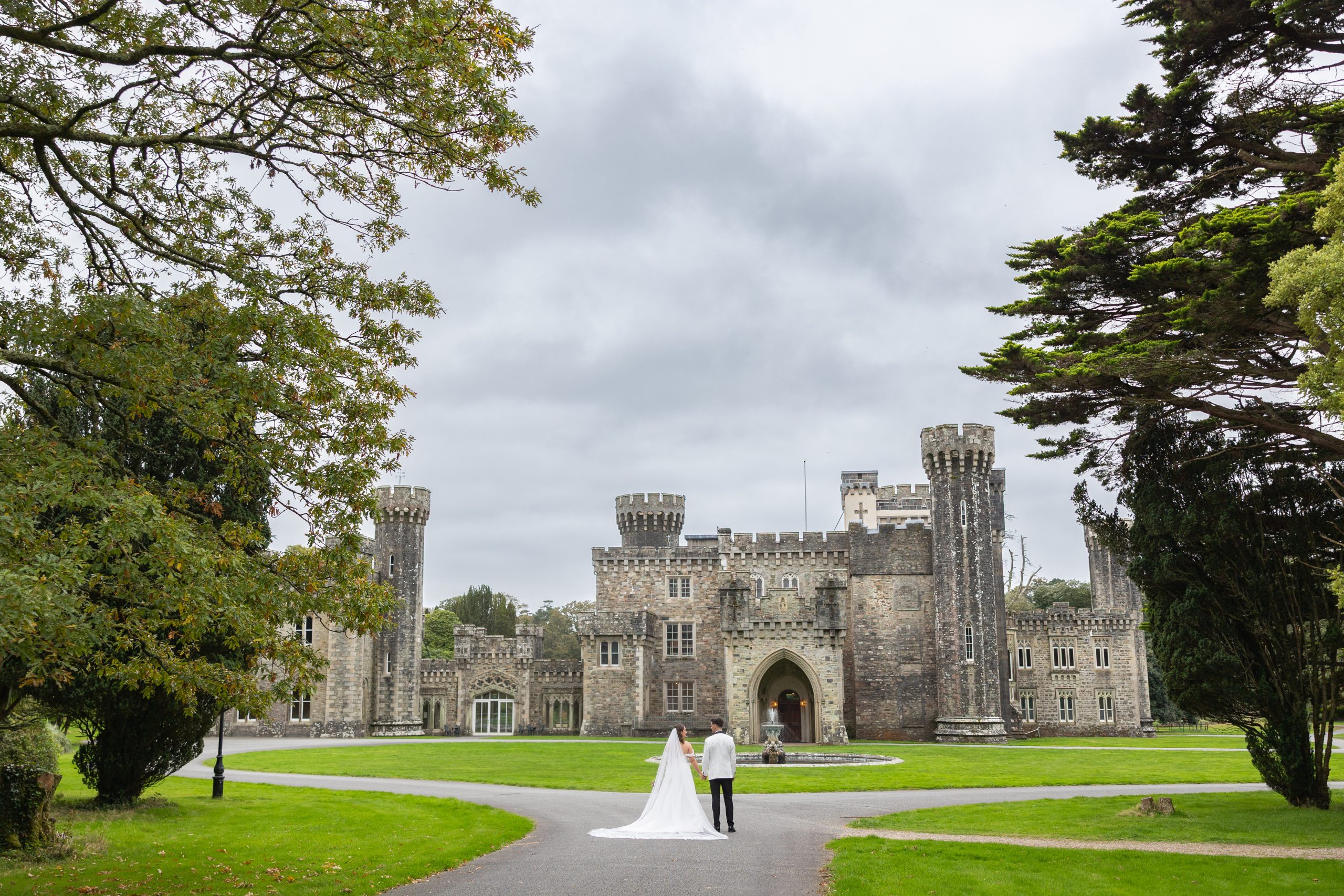 The bride and groom are standing on the lawn in front of Johnstown Castle, seen from behind. The castle forms a majestic backdrop, and the couple appears serene and elegant as they face the historic building.