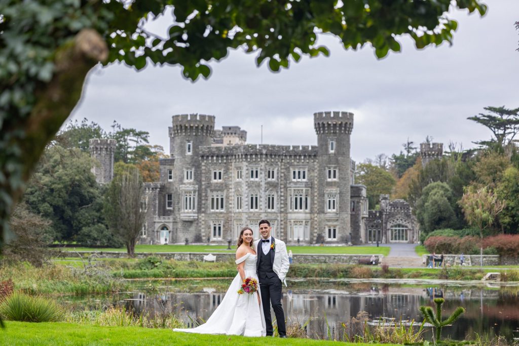 The bride and groom face the camera, standing on the lawn with the castle and its lake as a scenic backdrop, creating a romantic and picturesque setting