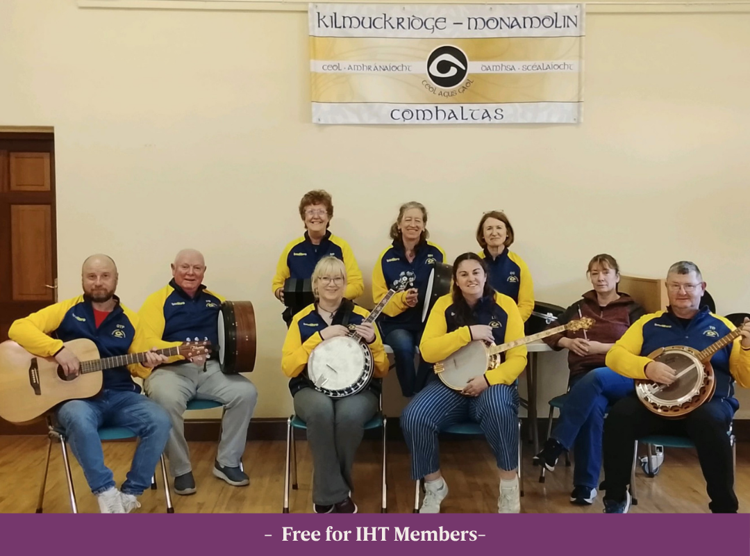 Group photo of the Kilmuckridge Monamolin Comhaltas Ceoltóirí Éireann (CCE) group, standing together in front of their flag in the background, holding their musical instruments.