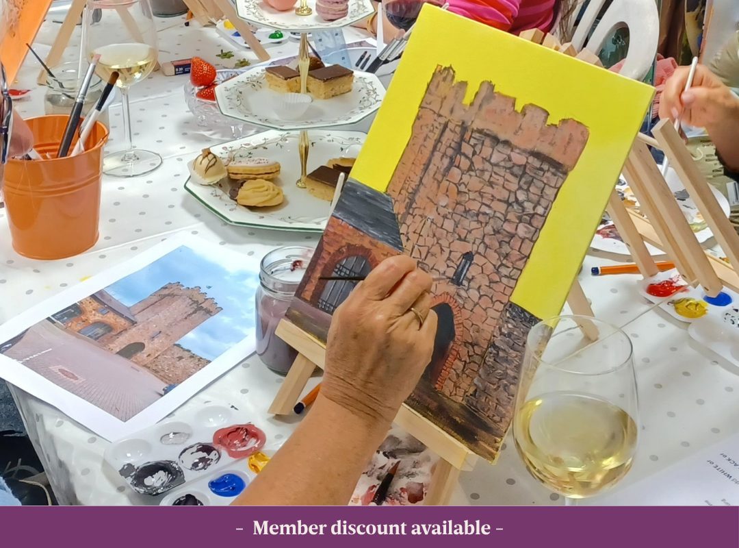 Artist painting an image based on an image of a castle with glasses on wine on the table.