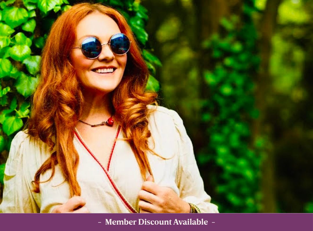 Summer of Love Jazz Web Image Sharon Clancy with bright red hair wearing sunglasses and a summer dress, standing outdoors in front of lush green foliage.